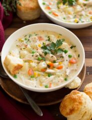Two bowls of chicken pot pie soup in white serving dishes set over wooden a wooden tabletop. Biscuits are served to the side.