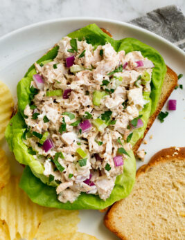 Tuna salad atop bread slices and lettuce leaves. Shown on a white plate with a side of potato chips.