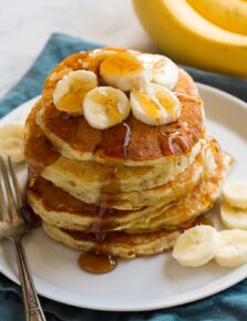 Stack of banana pancakes on a plate. Pancakes are topped with banana slices and maple syrup.