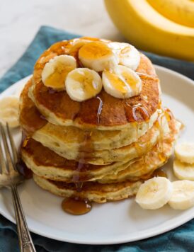 Stack of banana pancakes on a plate. Pancakes are topped with banana slices and maple syrup.