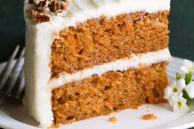Slice of carrot cake with cream cheese frosting on a white dessert plate sitting on a green cloth napkin.