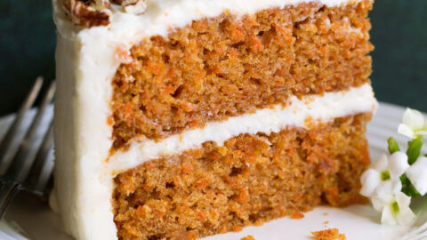 Slice of carrot cake with cream cheese frosting on a white dessert plate sitting on a green cloth napkin.
