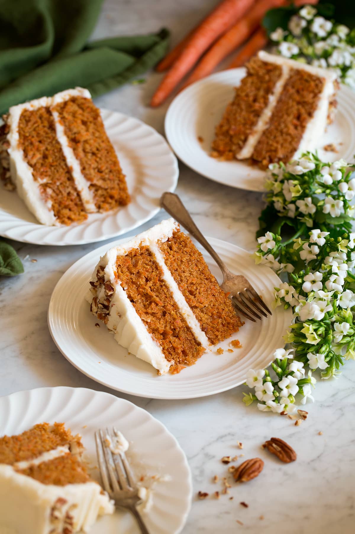 Four slices of carrot cake shown on white scalloped dessert plates with flowers and carrots around plates.