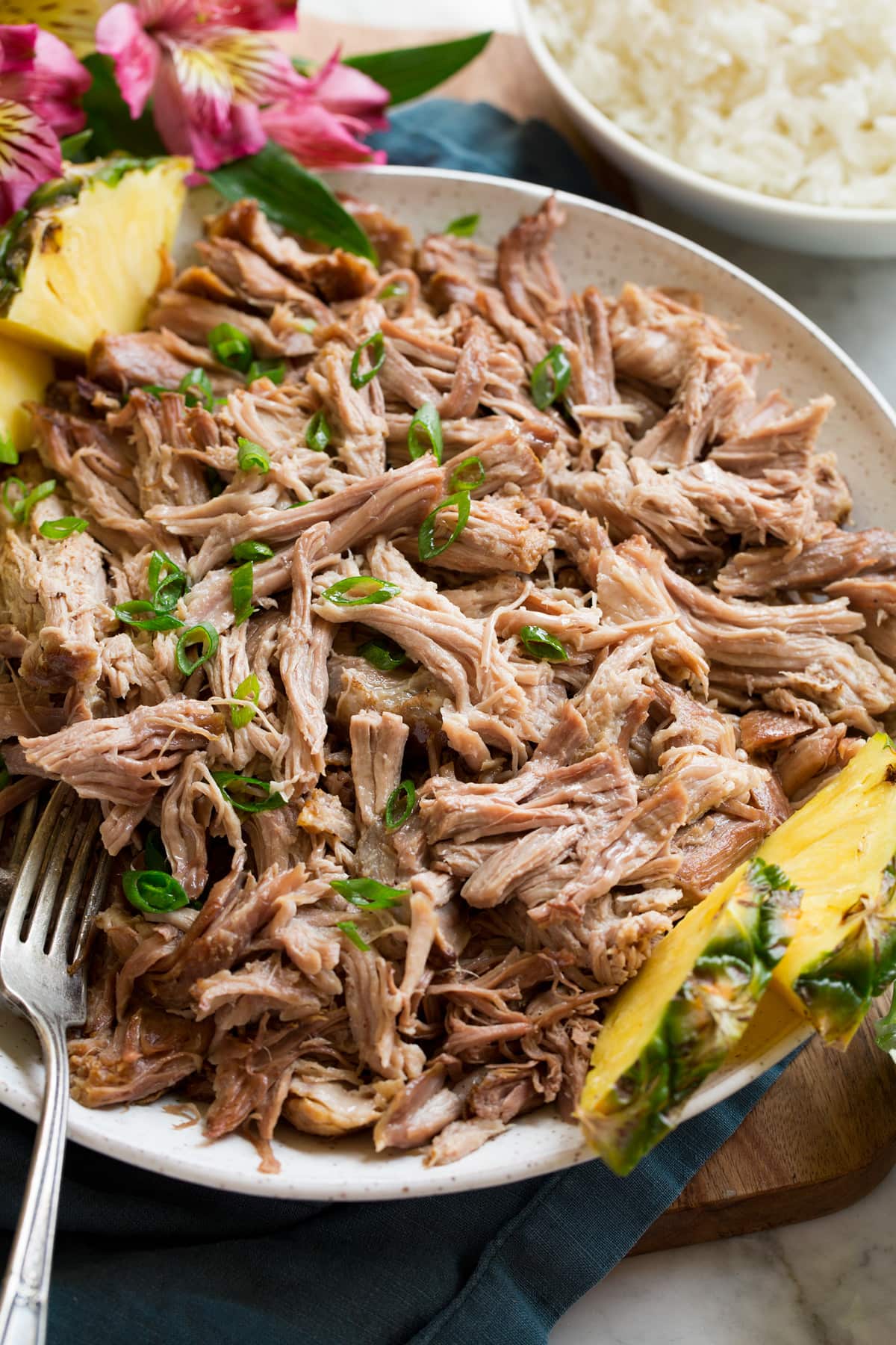 Shredded kalua pork shown from the side in a white serving bowl set over a blue napkin and wooden platter. Pineapple slices and tropical flowers are shown to the side.