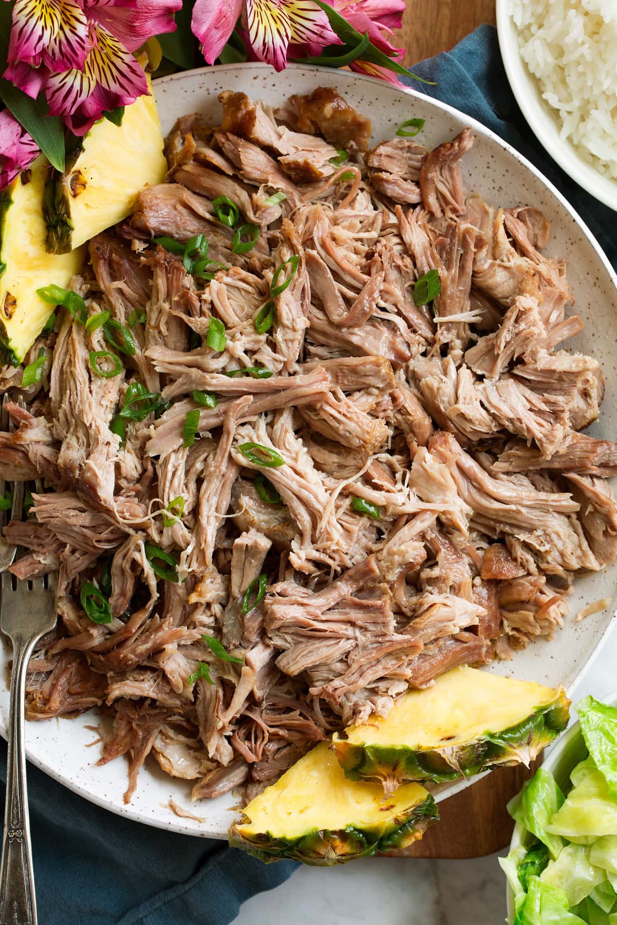 Overhead image of shredded Kalua pork in a white serving bowl with pineapple. It is garnished with sliced green onions.