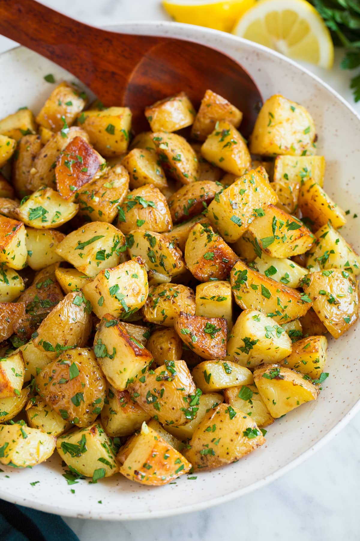 Bowl of roasted potatoes with lemon and parsley, wooden spoon is resting in potatoes.