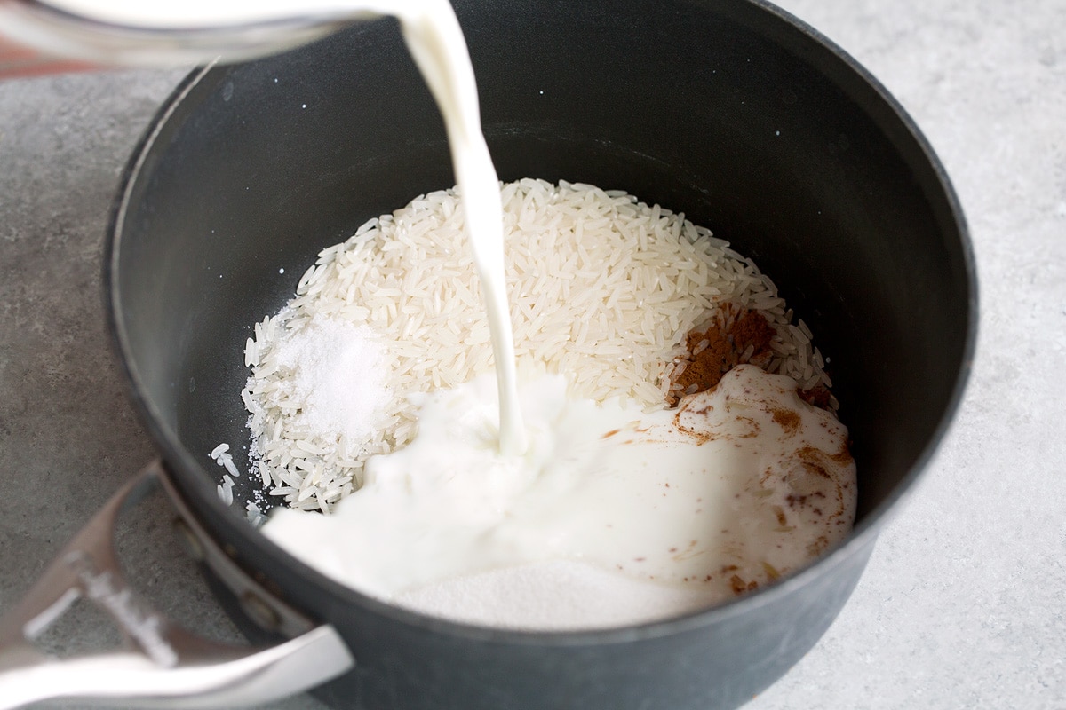 Milk being poured into a saucepan with rice, cinnamon, sugar and salt.