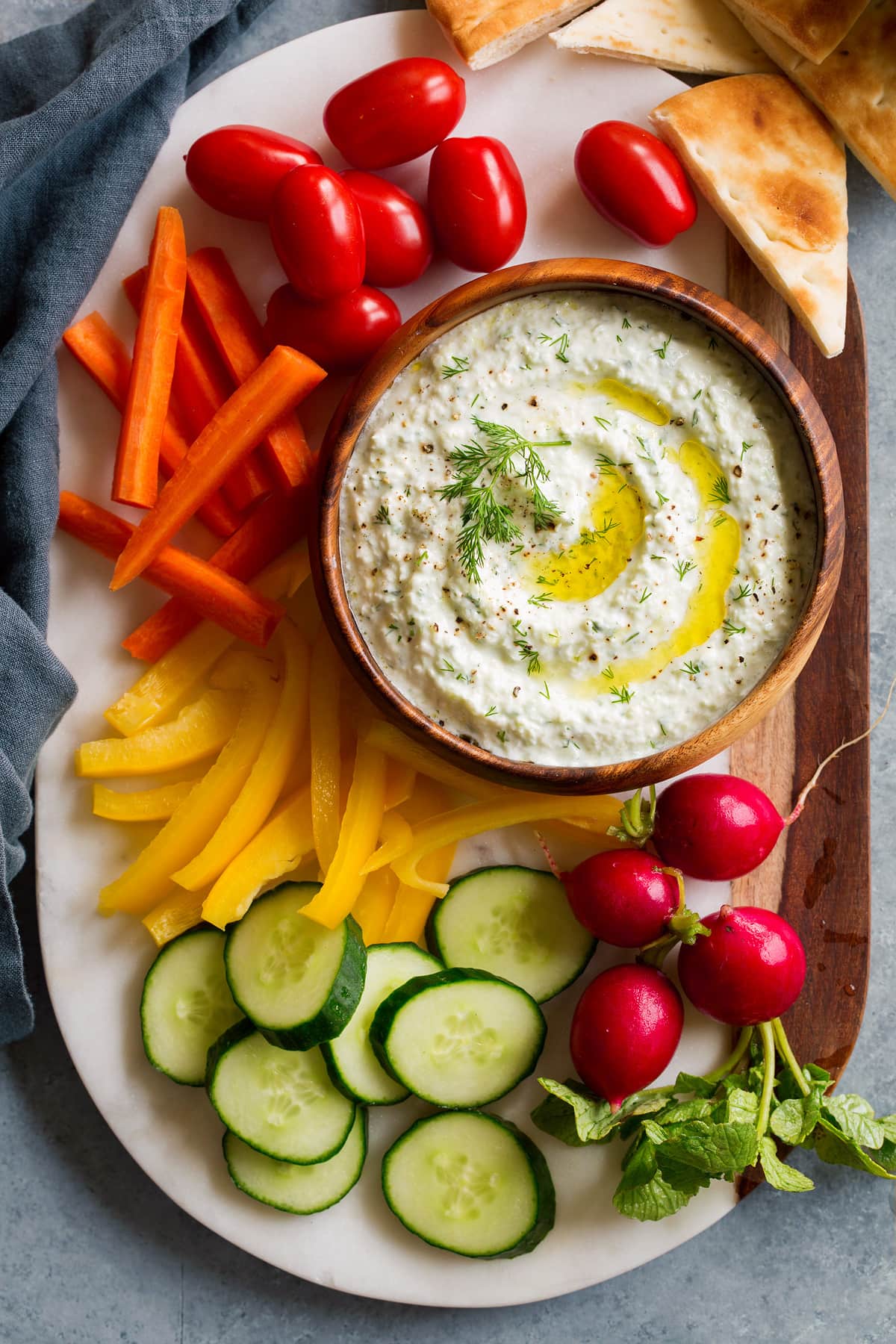 Overhead image of tzatziki sauce dip on a marble platter with fresh vegetables around it.