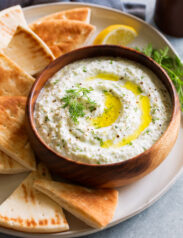 Bowl of homemade tzatziki sauce in wooden bowl on a platter with pita bread to the side.