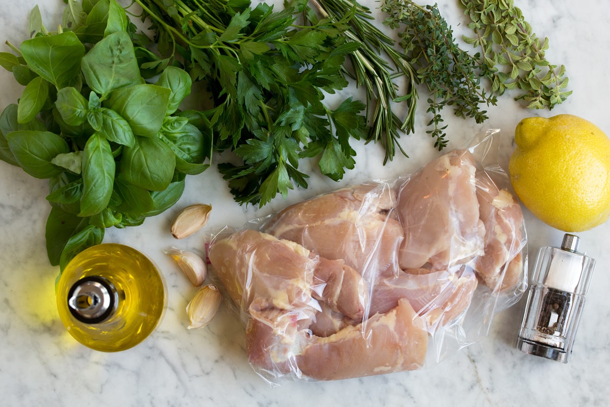 Image of ingredients used to make garlic herb grilled chicken and marinade. Includes olive oil, fresh basil, parsley, rosemary, thyme, oregano, garlic, lemon, salt and pepper.