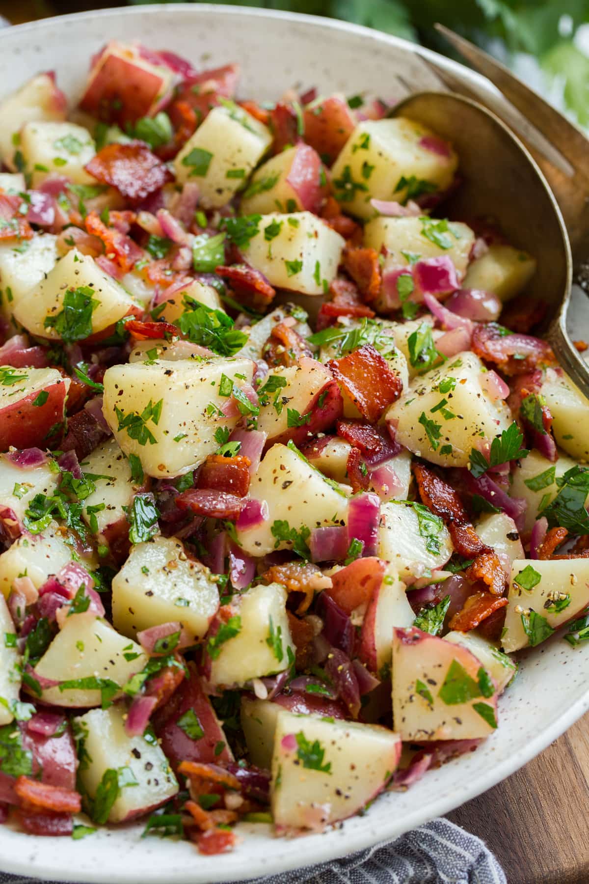 Close up image of german potato salad with red potatoes, parsley, bacon and onions in white ceramic serving bowl.
