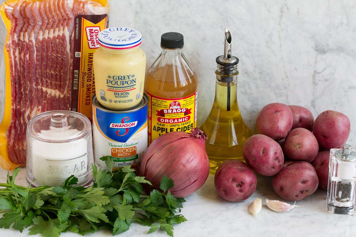 Image of ingredients that are used to make German potato salad. Includes red potatoes, olive oil, bacon, vinegar, onion, chicken broth, garlic, dijon mustard, parsley, and bacon.