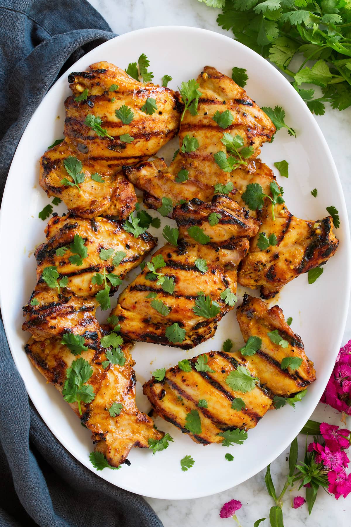 Thai Coconut Grilled Chicken Thighs shown overhead on a white oval platter. Chicken is garnished with cilantro and flowers are shown to the side.