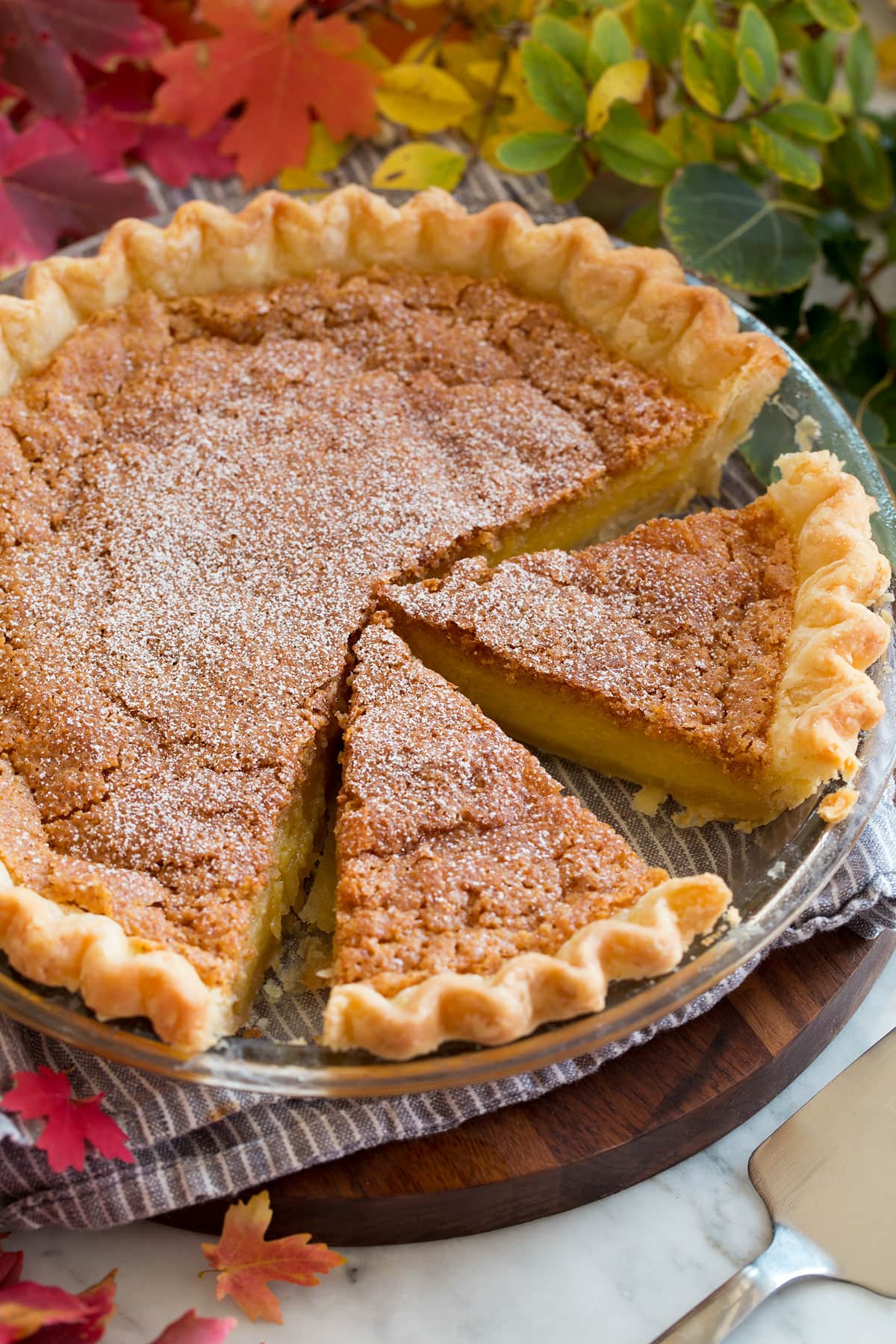 Image of chess pie cut into slices. Pie is shown in a glass baking dish set over a grey cloth on a wooden serving tray.