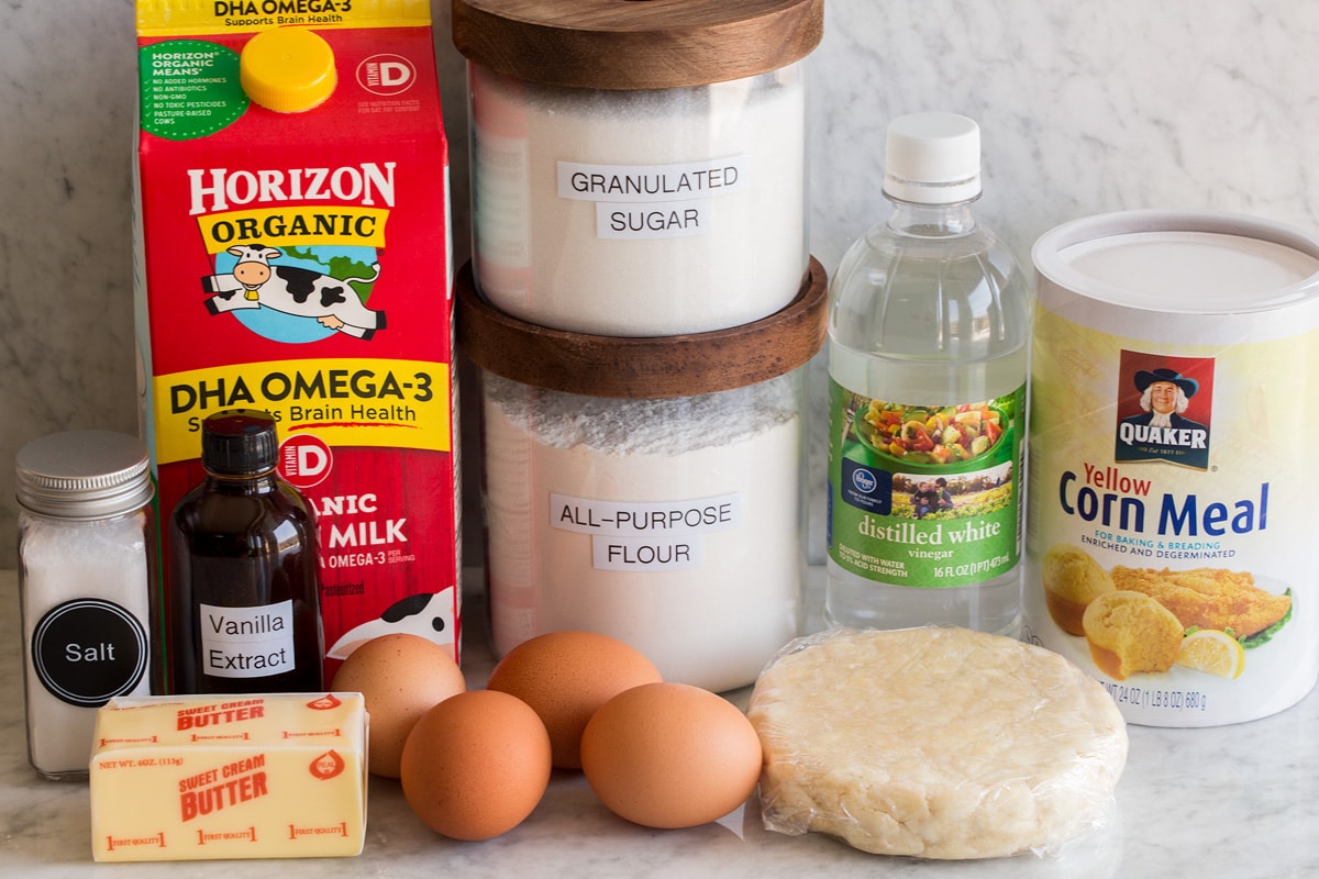 Image of ingredients used to make chess pie. Includes pie crust dough, cornmeal, vinegar, eggs, sugar, flour, milk, vanilla, butter and salt. 