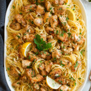 Chicken scampi shown on an oval serving platter. Includes thin spaghetti, lemon, browned chicken pieces and parsley.