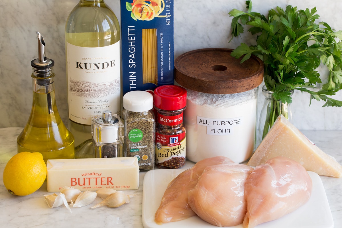 Image of ingredients used to make chicken scampi. Includes chicken breasts, olive oil, white wine, butter, garlic, lemon, pasta, Italian seasoning, red pepper flakes, salt, pepper, flour, parmesan and parsley.