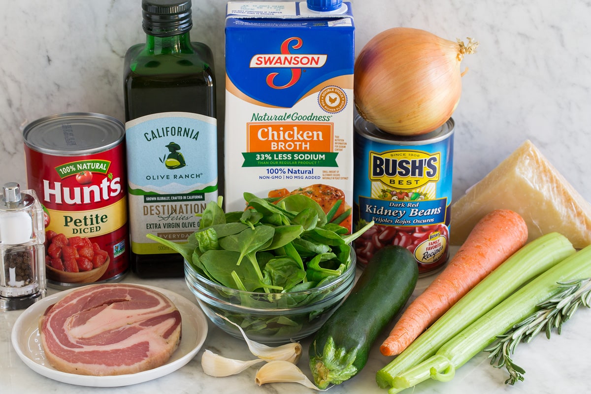 Image of ingredients used to make Minestrone soup. Shows celery, carrots, rosemary, zucchini, parmesan, kidney beans, yellow onion, spinach, chicken broth, garlic, pancetta, olive oil, canned tomatoes, salt and pepper.