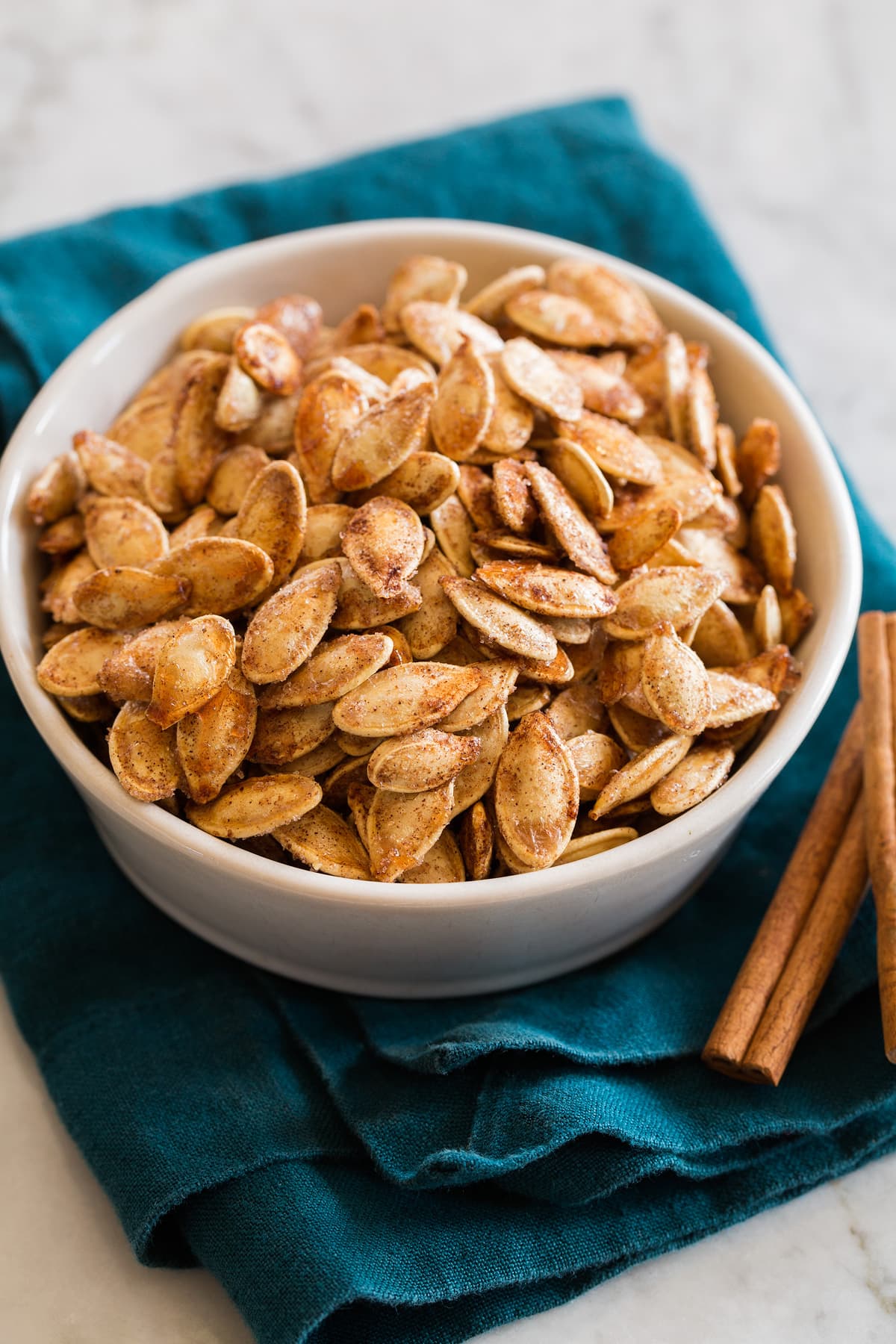 Image of cinnamon sugar roasted pumpkin seeds in a white bowl set over a blue cloth with cinnamon sticks shown to the side.