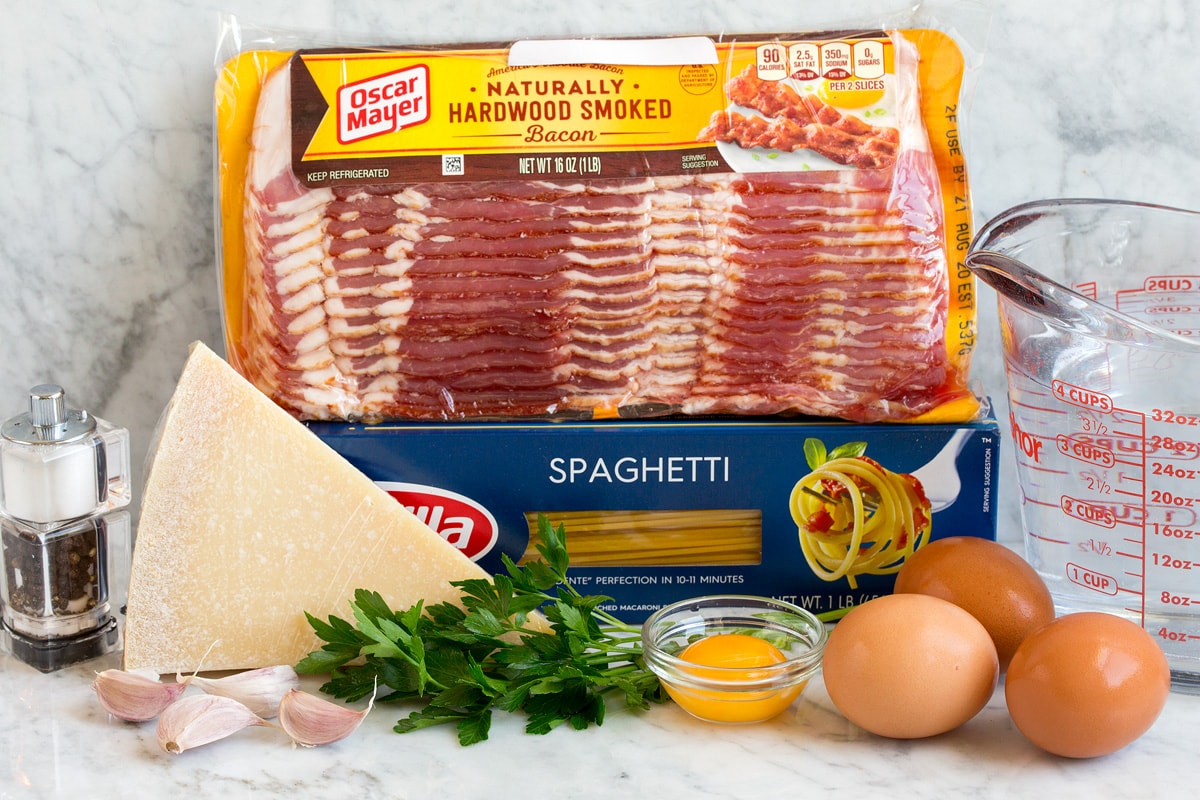 Image of pasta carbonara ingredients. Includes spaghetti, bacon, parsley, parmesan, eggs, egg yolk, water, garlic, salt and pepper.
