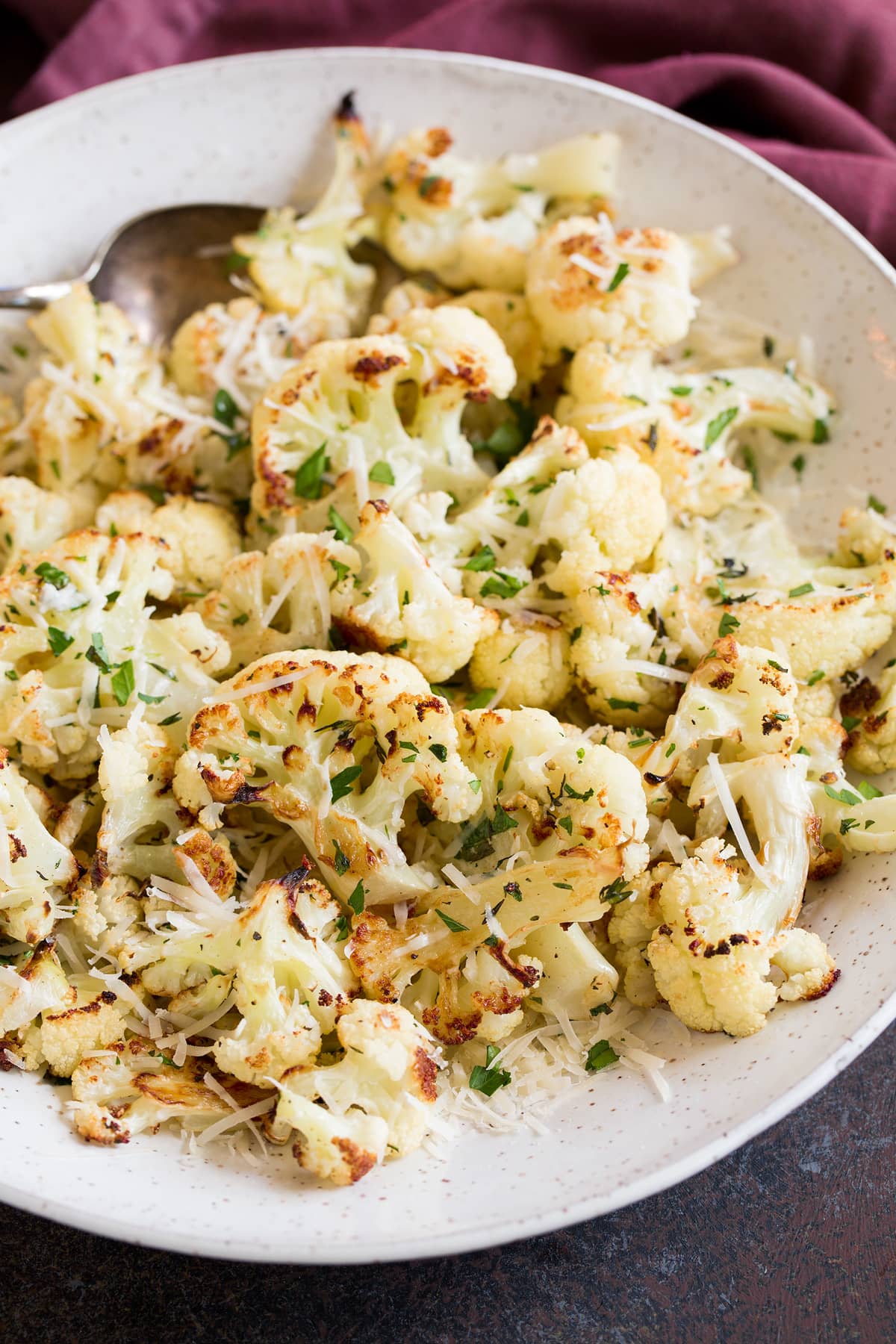 Roasted Cauliflower florets shown with caramelized edges. Served in a white bowl, cauliflower is garnished with herbs, garlic and parmesan.