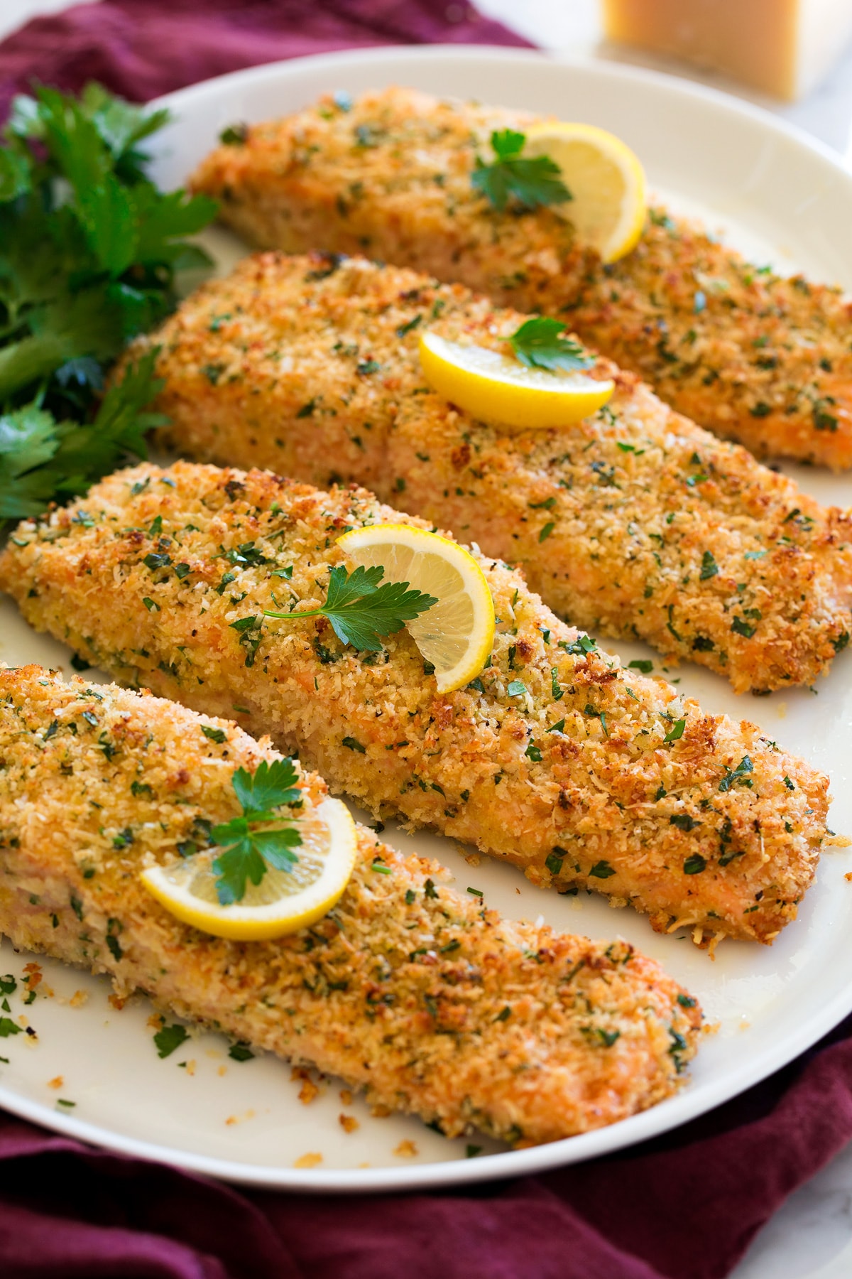 Four fillets of baked Parmesan Crusted Salmon shown on a white oval serving platter garnished with lemon slices and parsley.