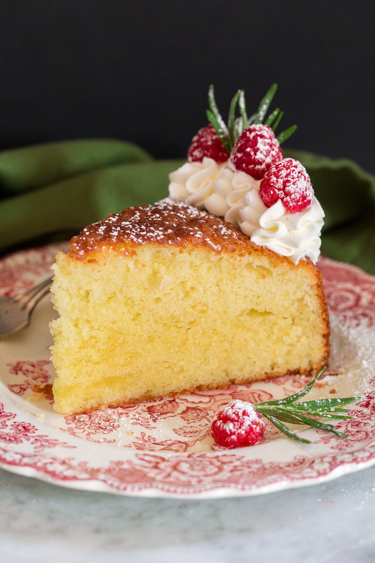 Photo of slice of olive oil cake decorated with whipped cream, raspberries and rosemary. Shown on a decorative red plate.