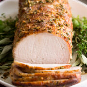 Pork Loin Roast served on a white platter with fresh herbs. Pork is shown sliced at the forefront.