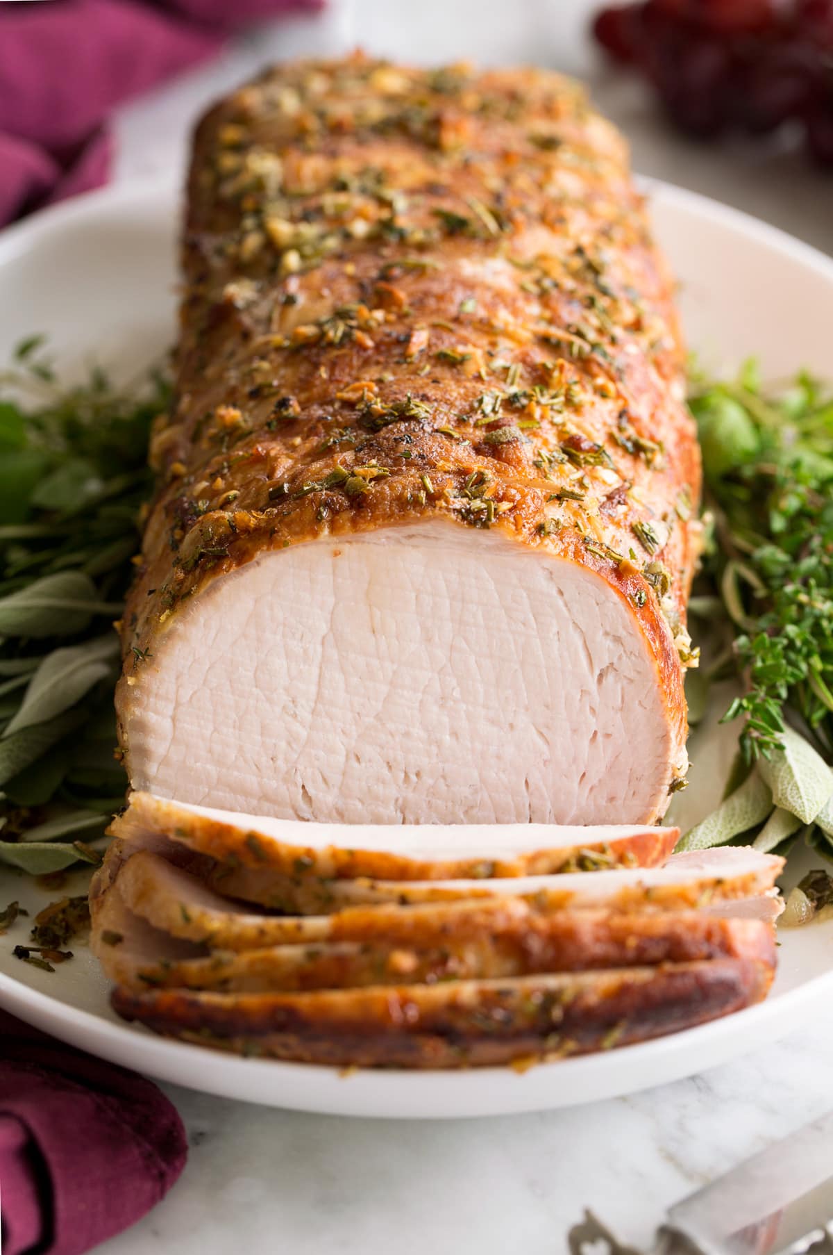 Pork Loin Roast served on a white platter with fresh herbs. Pork is shown sliced at the forefront. 