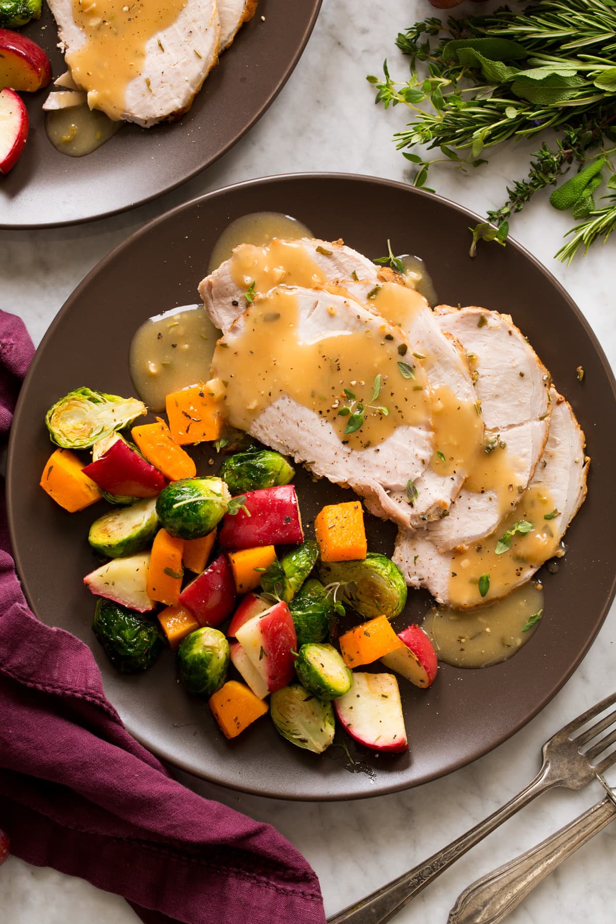 Pork loin shown cooked and sliced with gravy atop. Shown on a brown plate with a side of roasted vegetables and apples.