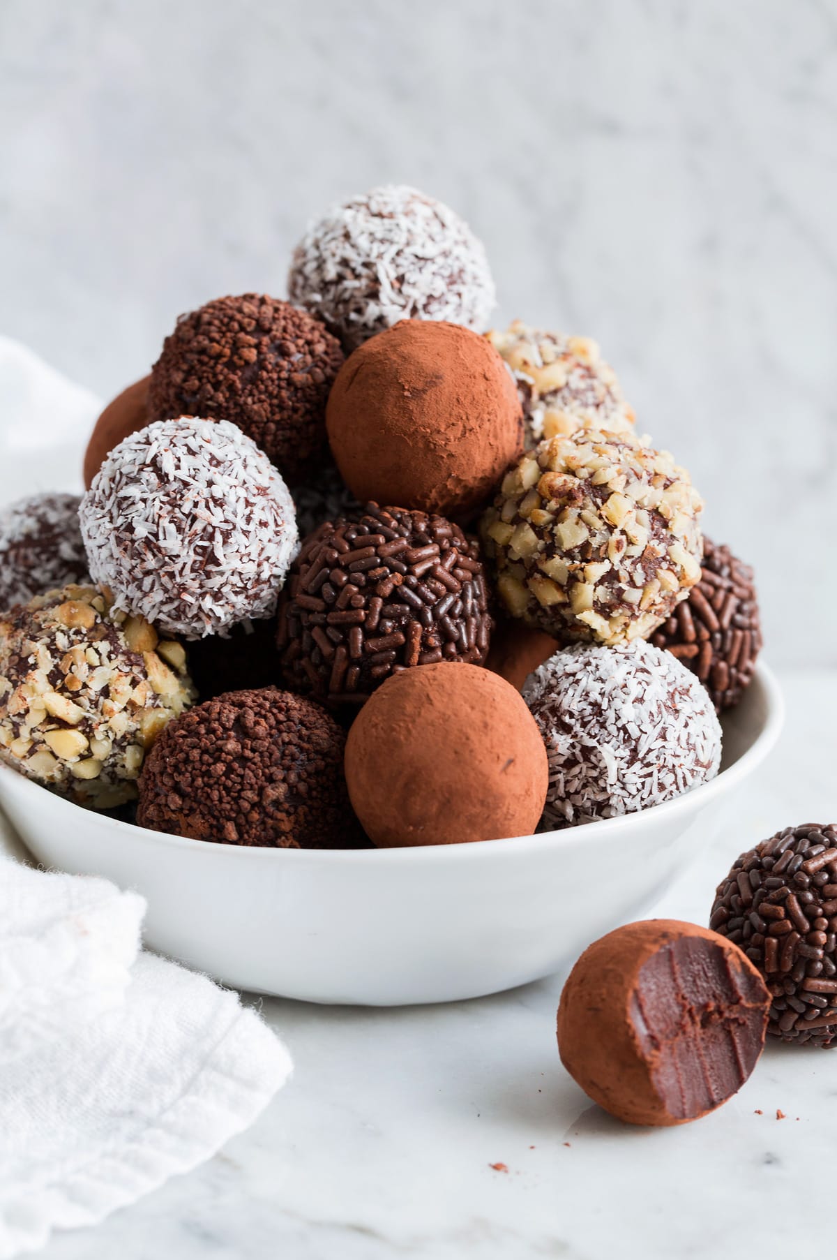 Stack of homemade chocolate truffles covered in cocoa, nuts, sprinkles and coconut piled into a pyramidal shape in a white bowl set over a marble surface.