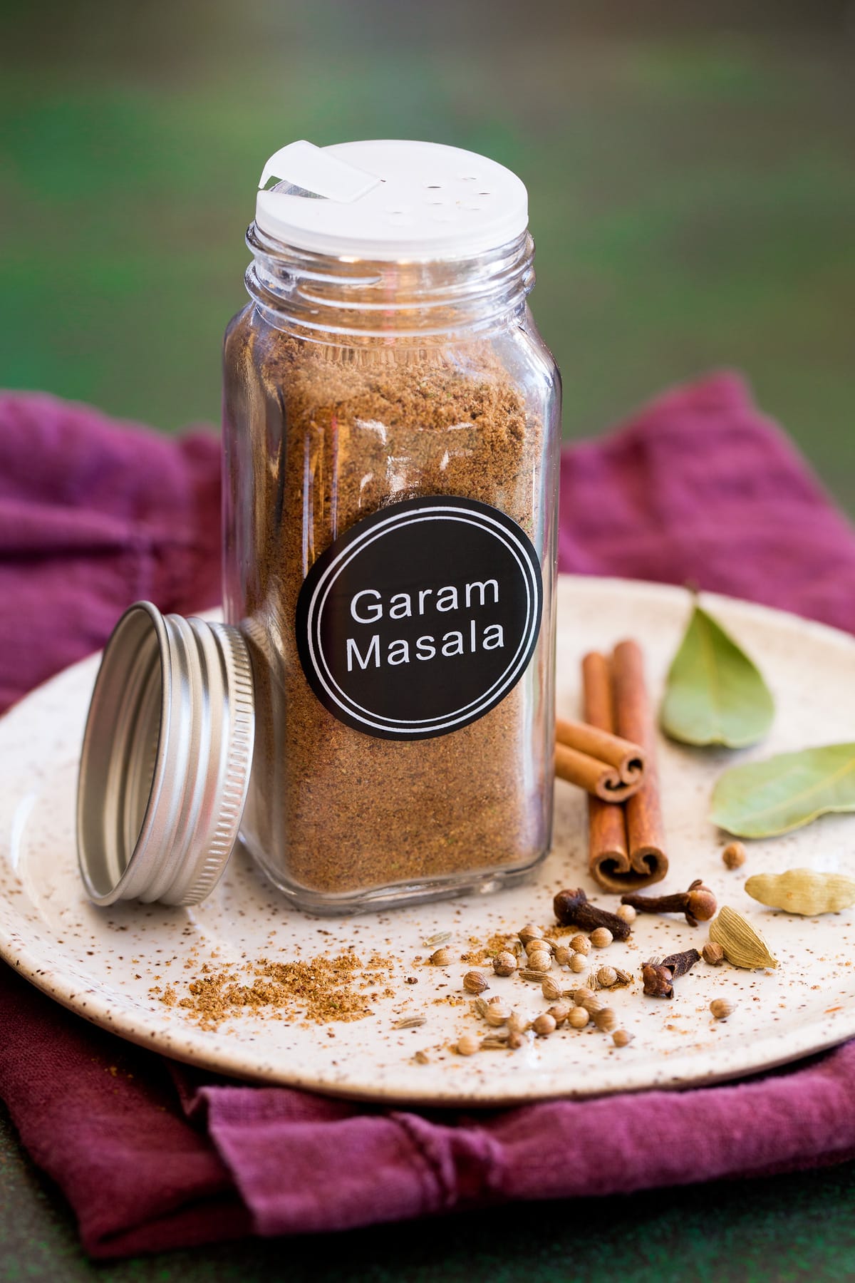 Garam masala spice blend in a glass jar set over a plate with whole spices next to it.