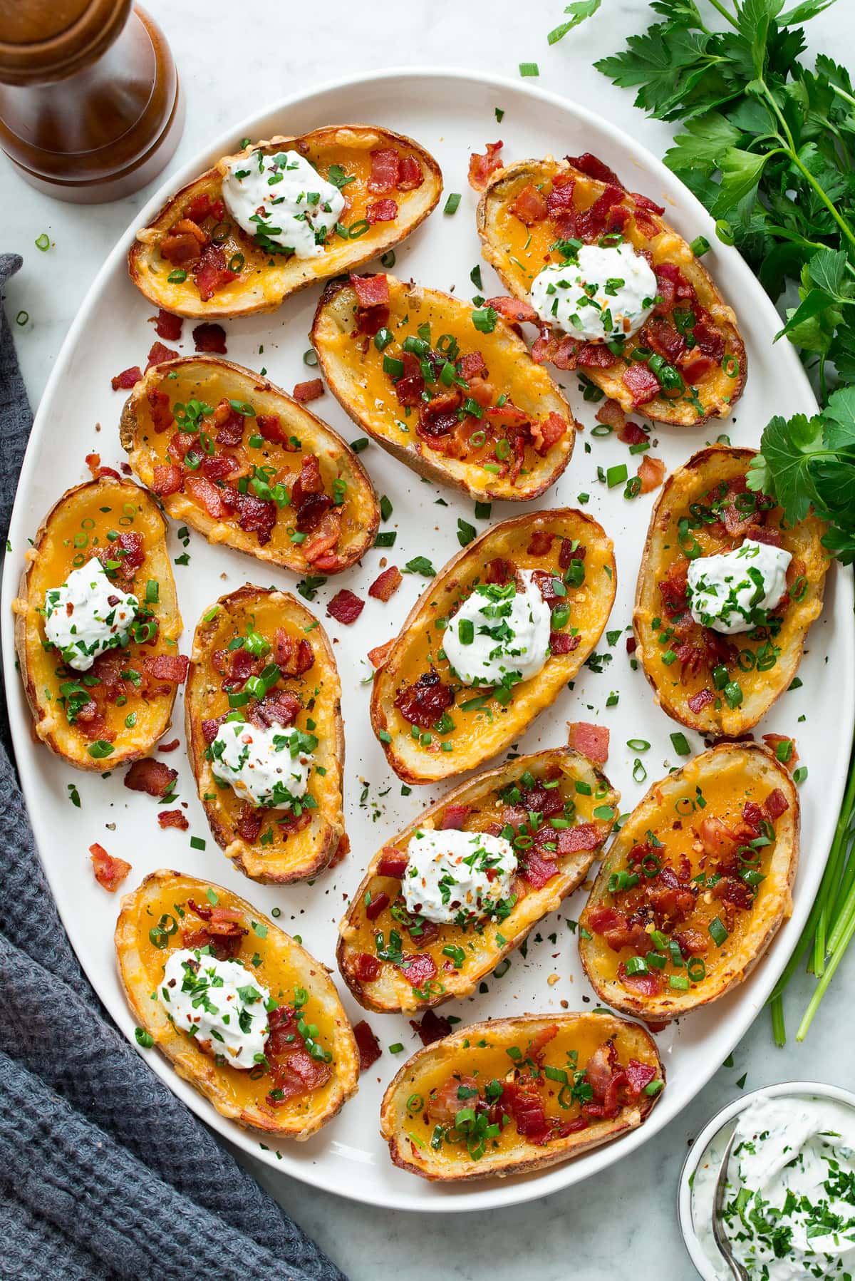 Potato skins on a white oval serving platter shown from above. Potato skins are garnished with green onions, bacon and topped with a dollop of herbed sour cream.