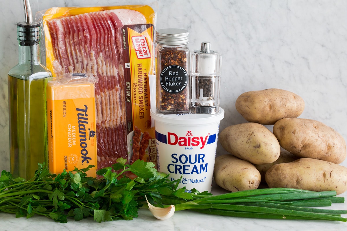 Photo of ingredients used to make potato skins. Includes russet potatoes, bacon, olive oil, cheddar cheese, sour cream, salt, pepper, red pepper flakes, parsley, green onions and garlic.