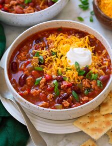 Serving of turkey chili in a white bowl set over a white plate with a spoon and saltine crackers to the side. Chili is topped with shredded cheddar cheese and sour cream.