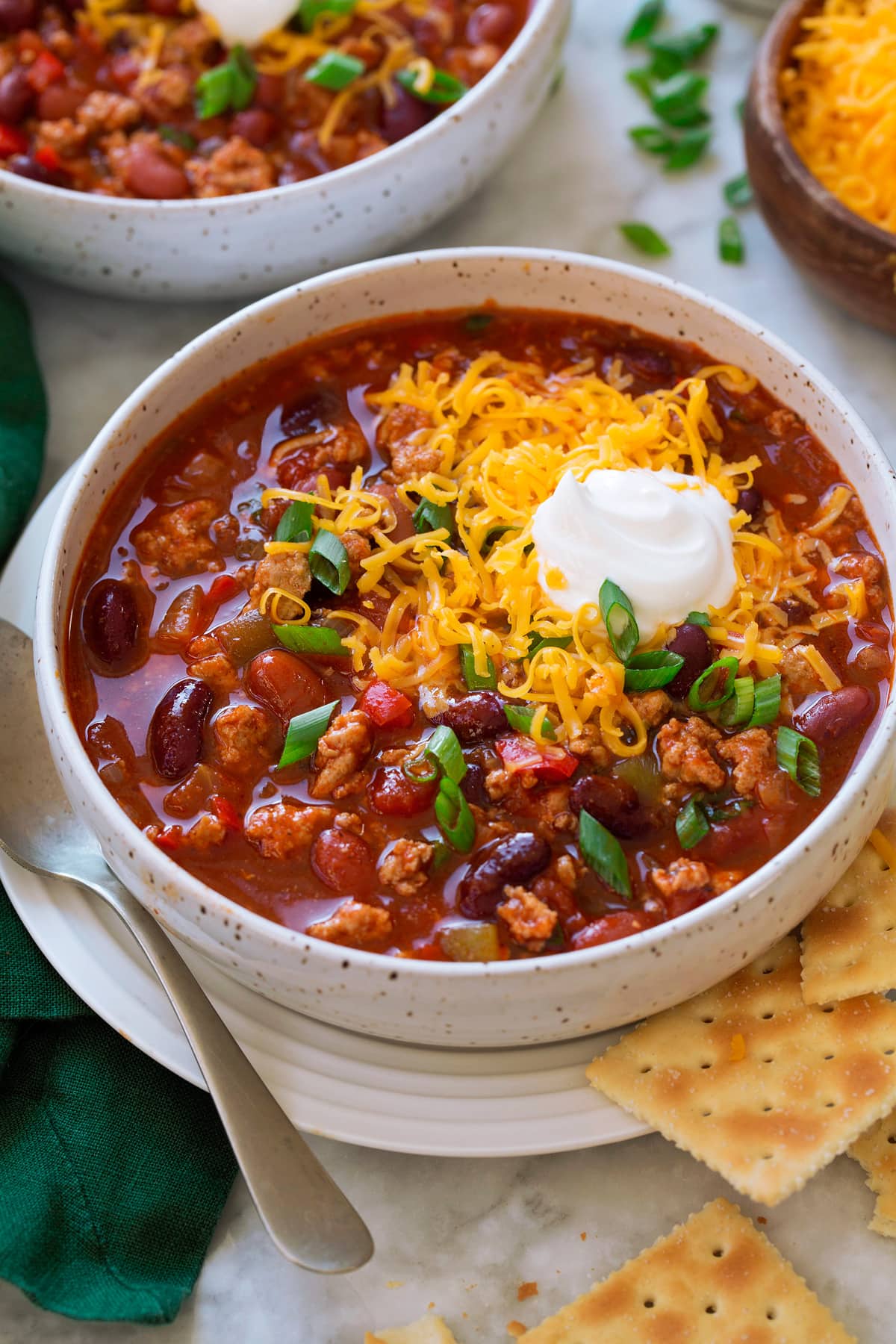 Serving of turkey chili in a white bowl set over a white plate with a spoon and saltine crackers to the side. Chili is topped with shredded cheddar cheese and sour cream.