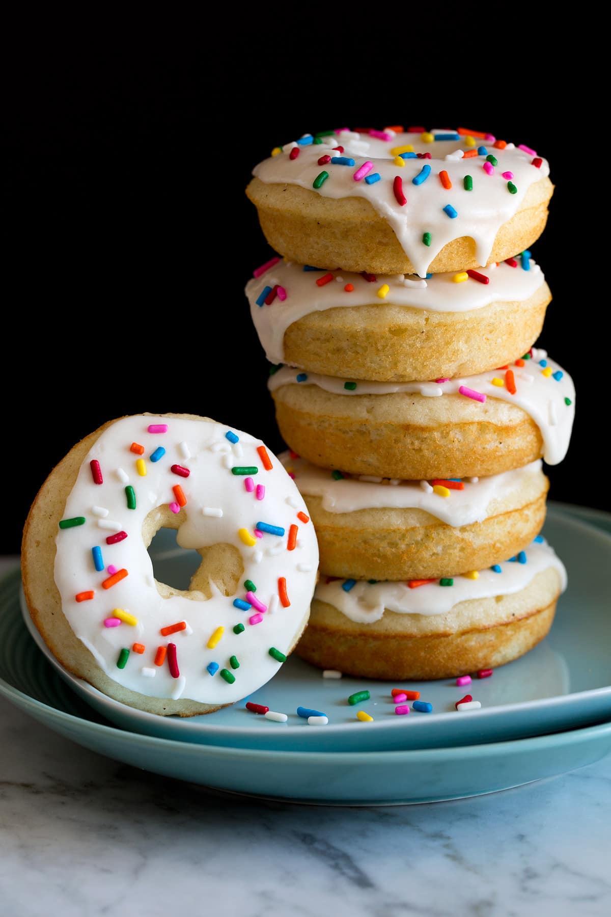 Image of stack of baked donuts on a plate. They are covered in icing and sprinkles.