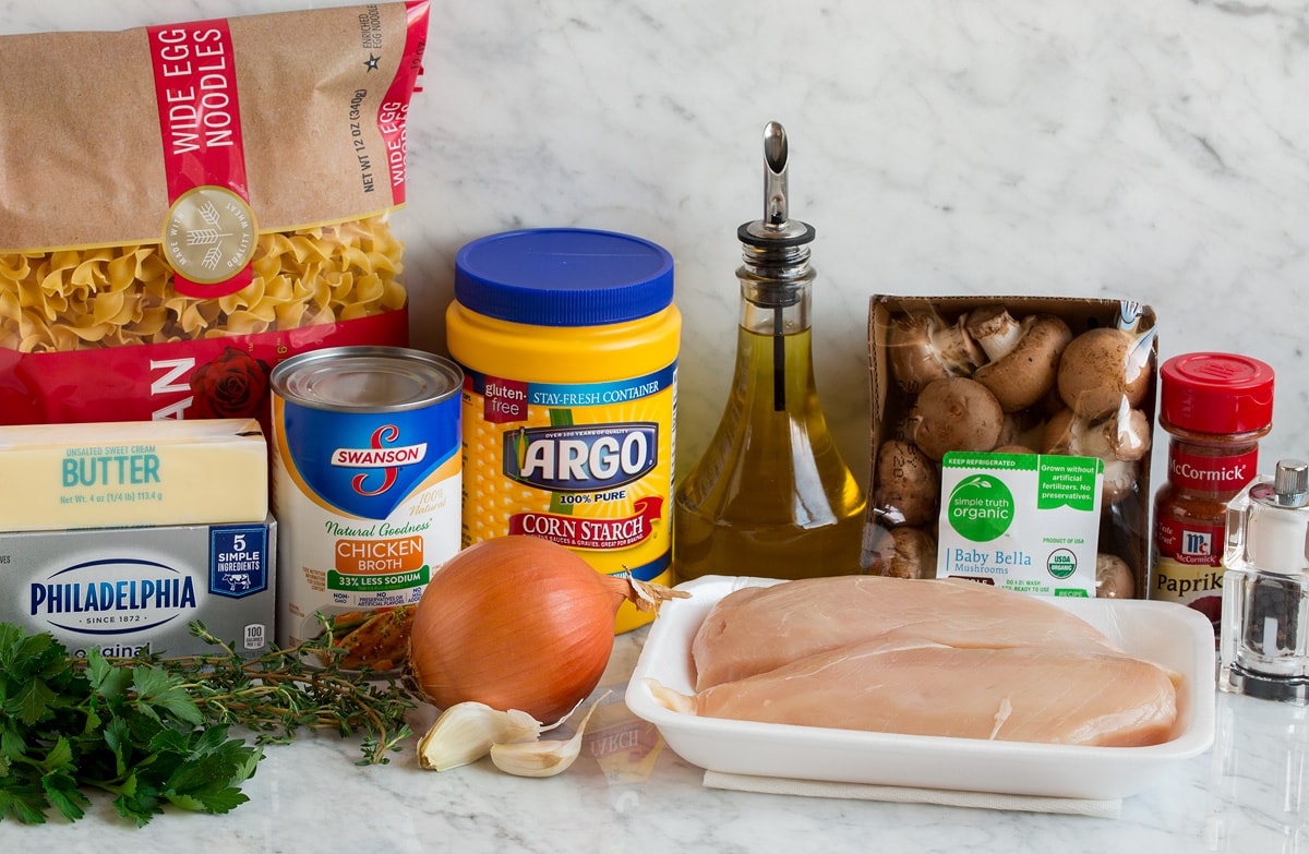 Photo of ingredients used to make chicken stroganoff. Includes chicken breasts, mushrooms, yellow onion, garlic, parsley, cream cheese, butter, chicken broth, thyme, egg noodles, olive oil, paprika, salt and pepper.