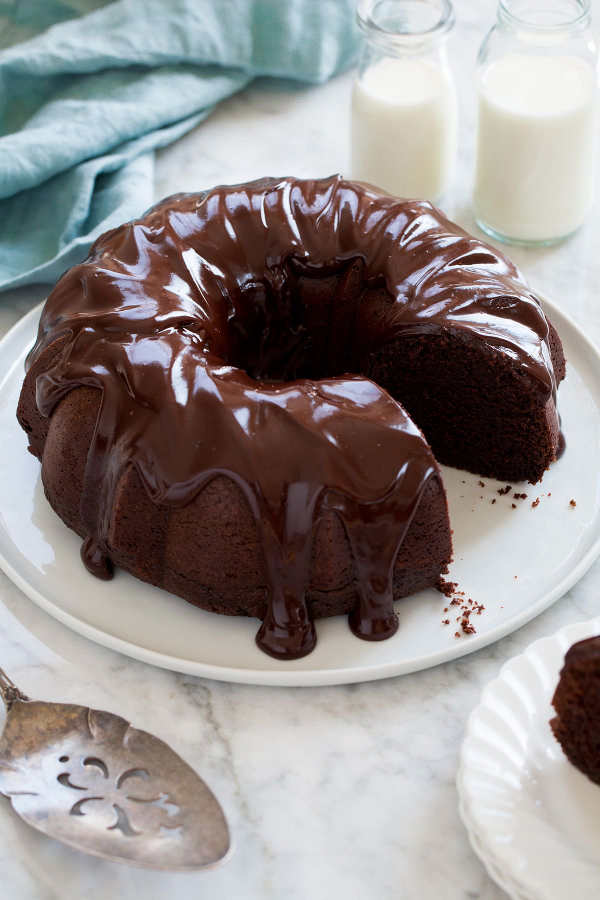 Whole chocolate bundt cake with one slice removed shown on a white platter with milk in the background and blue cloth.