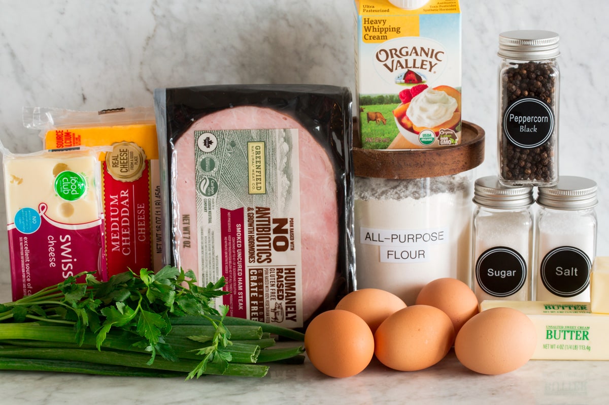 Photo of ingredients used to make a homemade quiche and crust. Includes flour, butter, salt, sugar, pepper, eggs, parsley, green onions, ham, cheddar and Swiss cheese.