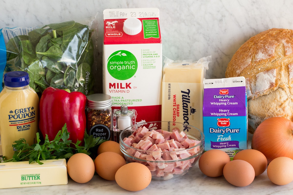 Photo of ingredients used to make a strata. Includes bread, eggs, onion, cream, cheddar cheese, milk, ham, red pepper flakes, bell pepper, spinach, dijon mustard and butter.