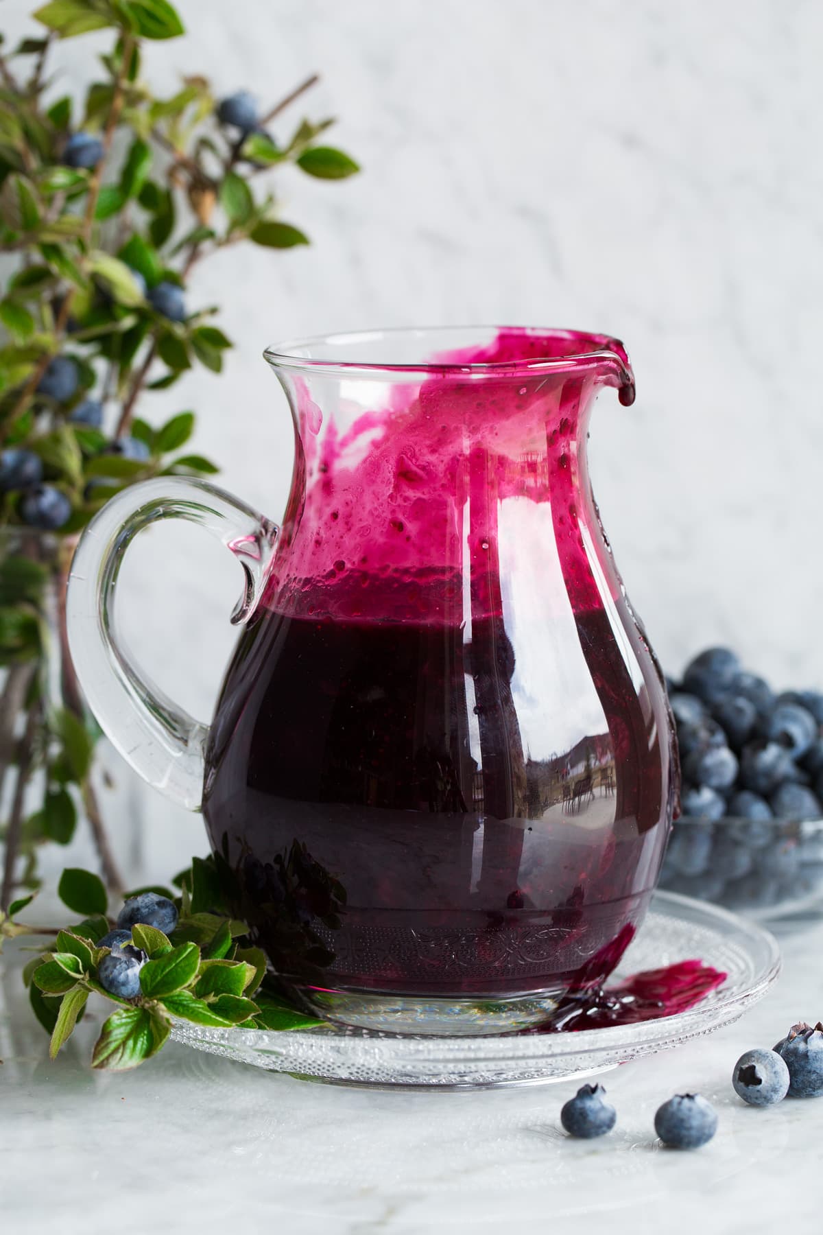 Blueberry syrup in a glass pitcher set over a glass plate.