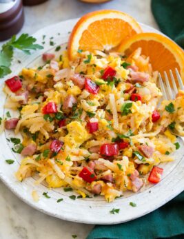Photo: Serving of breakfast hash on a white ceramic plate with oranges to the side. Hash is made with shredded hash browns, ham, eggs, bell pepper, parsley and onion.