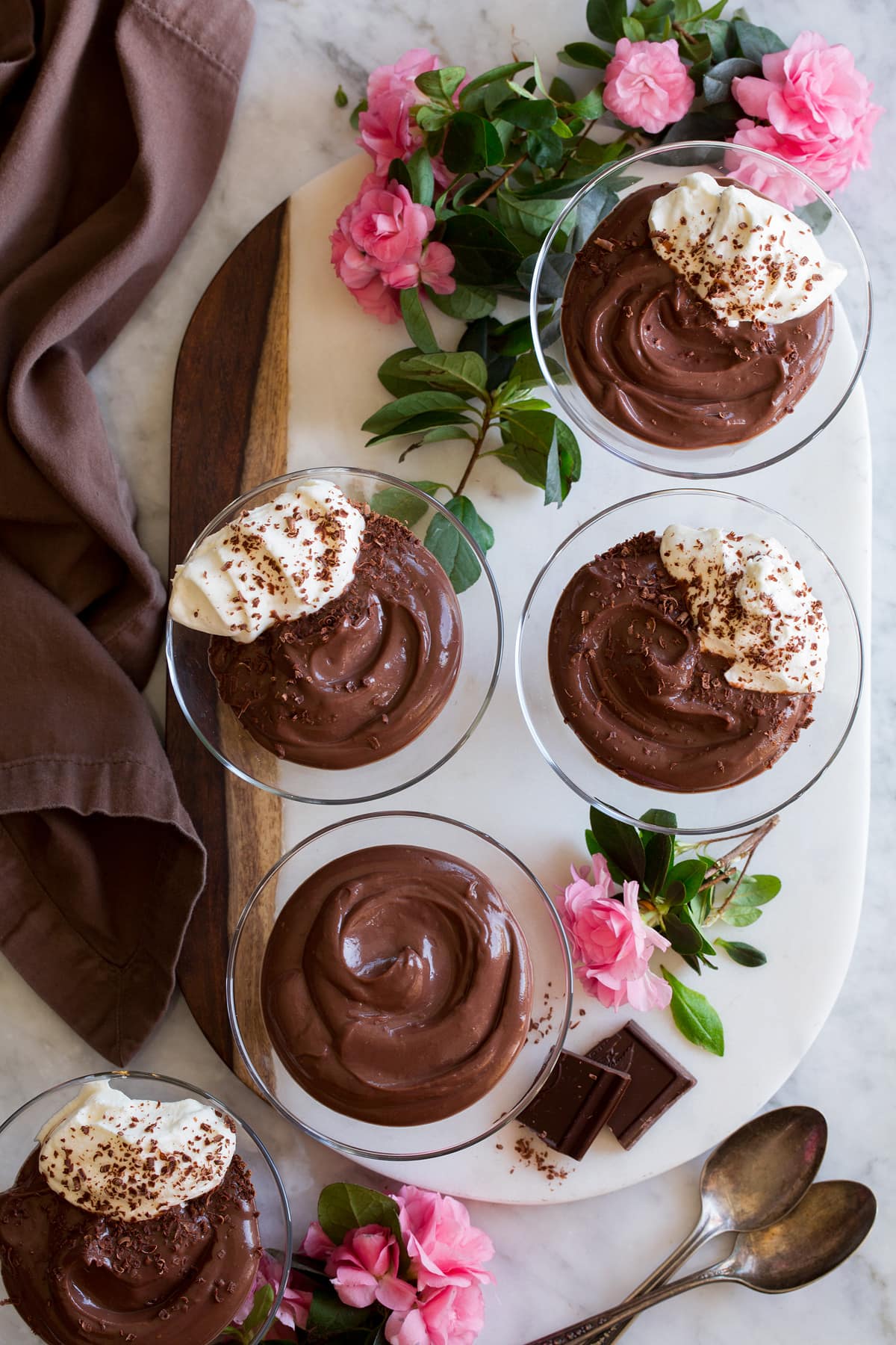 Overhead photo of 5 servings of chocolate pudding in glass cups resting on a marble surface with pink flowers and a brown cloth surrounding them.