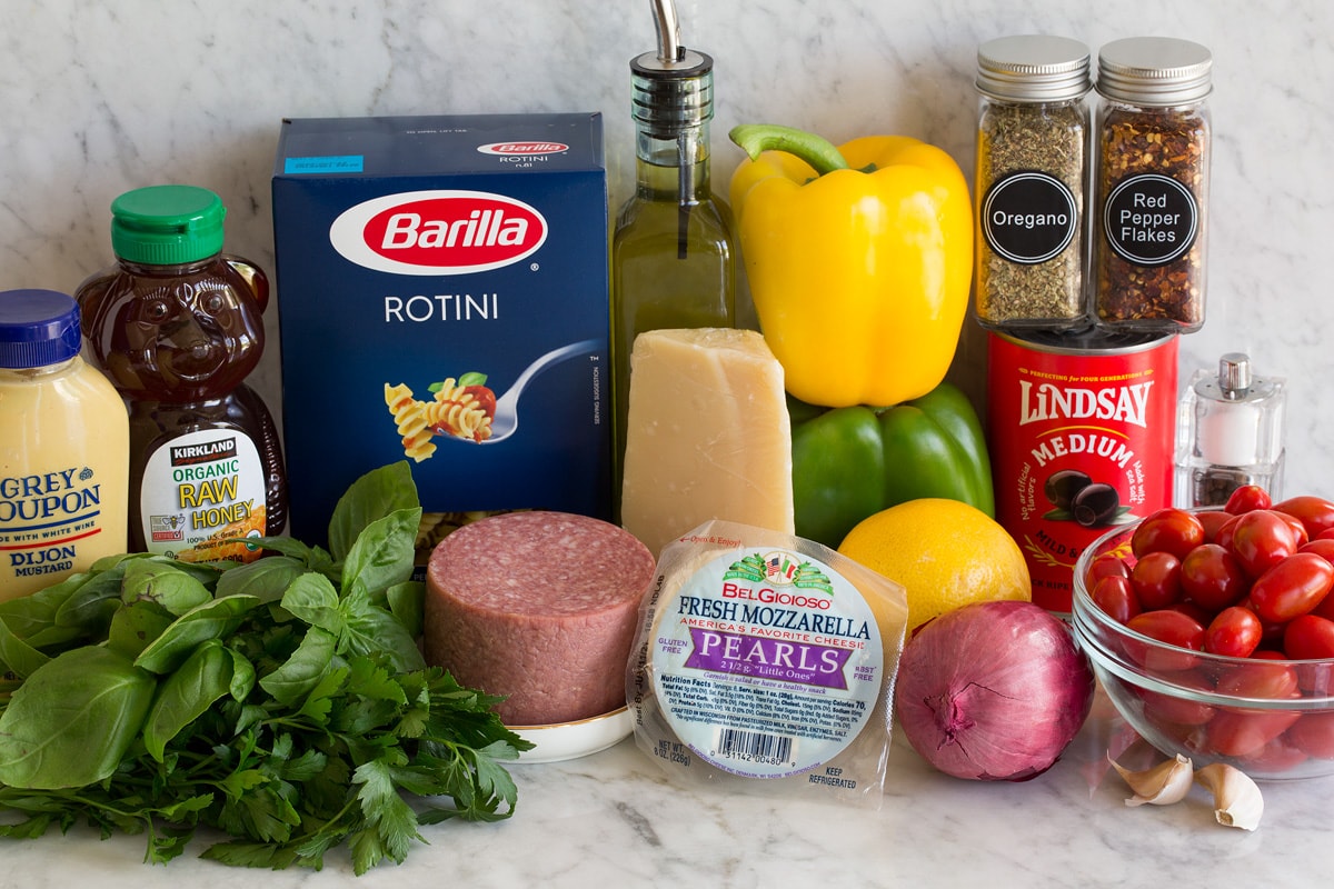 Photo: Ingredients displayed that are used to make Italian pasta salad. Includes rotini pasta, bell peppers, onion, lemon, parmesan, mozzarella, tomatoes, olives, herbs, garlic, salami, honey and dijon.
