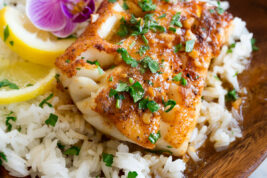 Four cod fillets shown overhead on a white oval platter. They are covered in a sauce and garnished with parsley. A package of butter and fresh parsley are shown to the side.