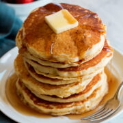 Stack of homemade pancakes on a white plate. Pancakes are topped with maple syrup and butter.