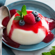 Photo: Single serving of a disk of Panna Cotta shown on a blue plate covered with raspberry sauce, fresh blueberries and mint.