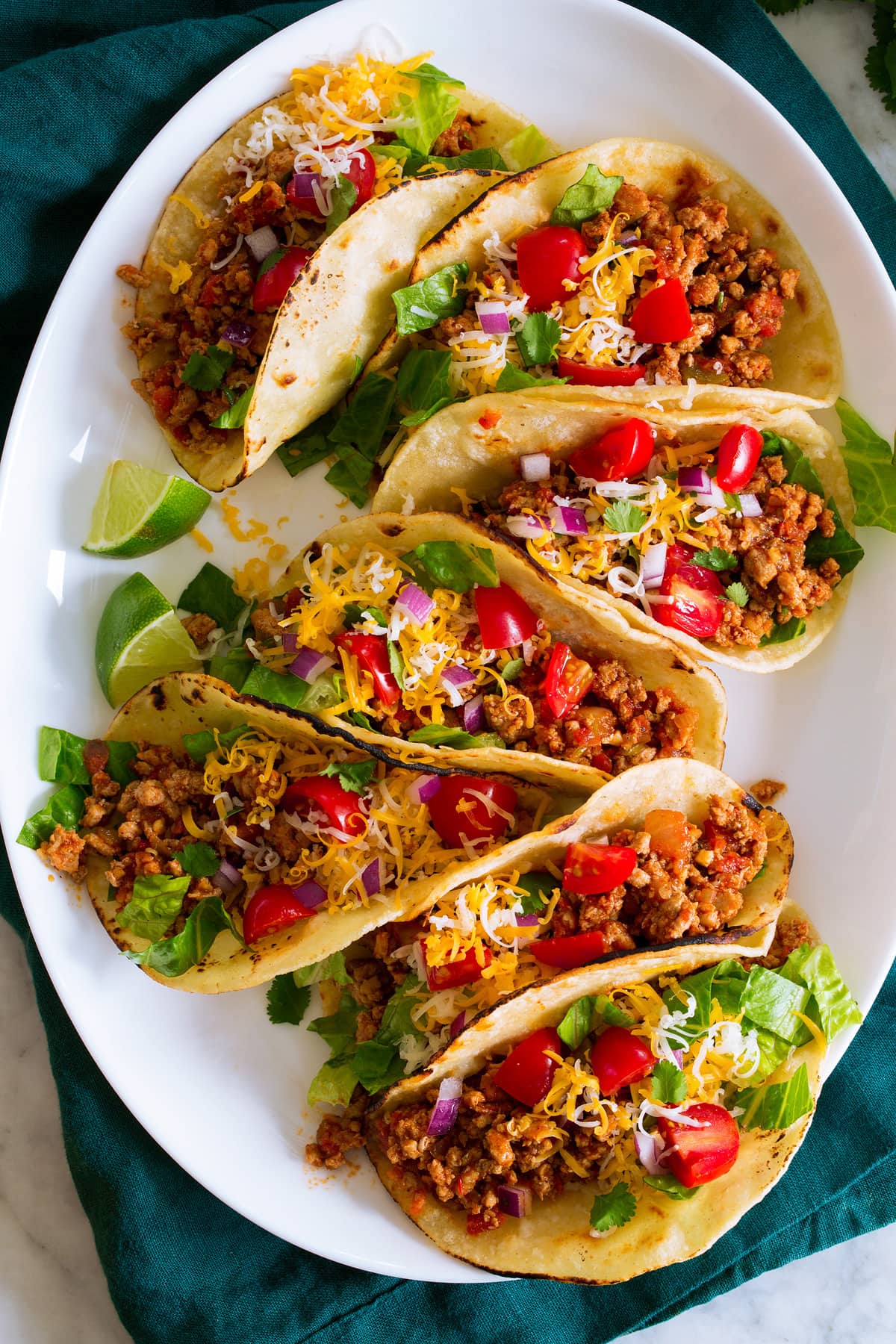 Photo: 7 ground turkey tacos in a row shown on a white oval platter from above. Tacos are filled with meat, tomatoes, lettuce, cheese and red onion.