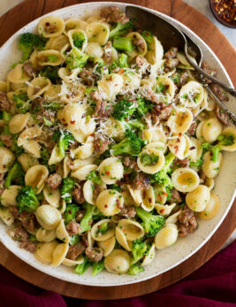 Photo: Orecchiette pasta with sausage and broccoli in a white serving bowl set over a wooden platter on a marble surface. It is shown from above.